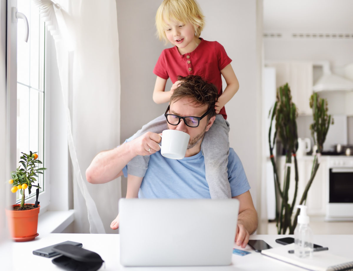 man working on a laptop at the kitchen table with toddler on his shoulders