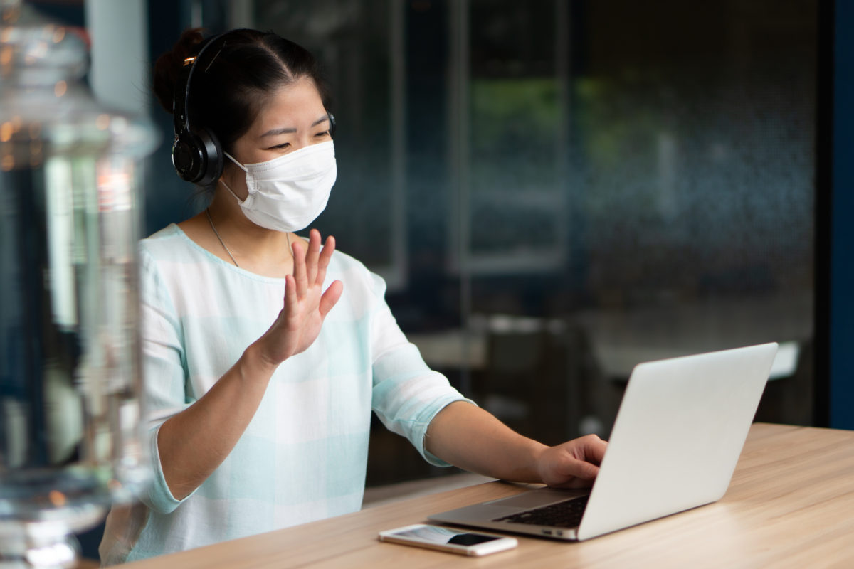 Young Asian woman wearing face mask and headphones and meeting with a remote coworker by using computer video call confernece