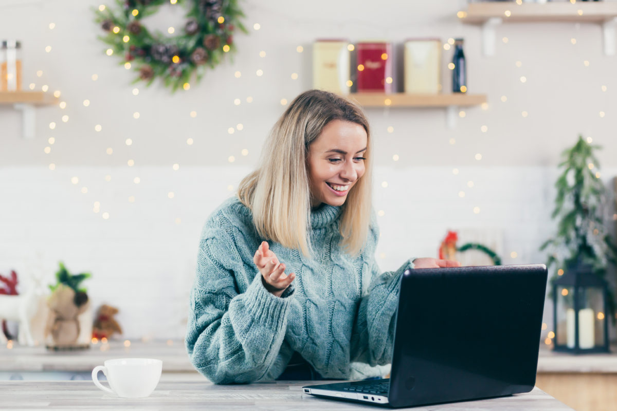 White woman in a winter swaeter with holiday decorations behind her participating in a virtual holiday party