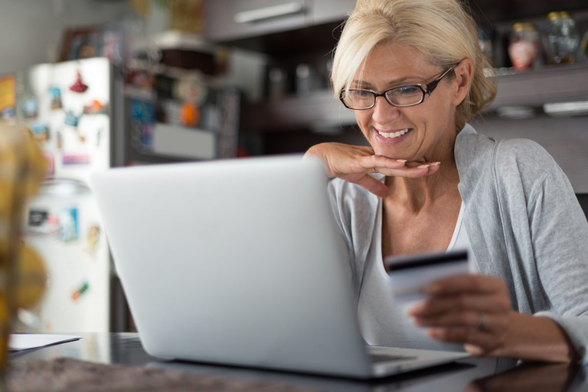White woman at her kitchen table, with her credit card in hand, participating in a virtual fundraising event on her laptop.